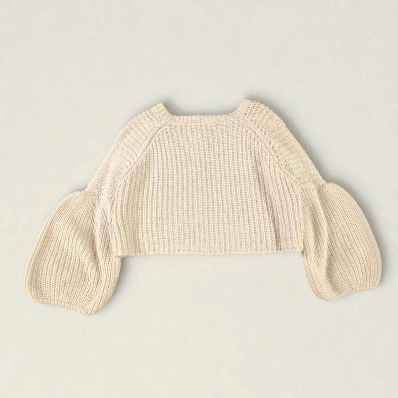 K&ouml;nnte beinhalten: Ein cremefarbener bauchfreier Pullover mit einer gerippten Strickstruktur am K&ouml;rper und an den &Auml;rmeln. Das Kleidungsst&uuml;ck hat einen weiten, runden Ausschnitt und volumin&ouml;se, bauschige &Auml;rmel, die an den B&uuml;ndchen schmaler zulaufen.