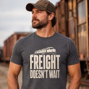 以下が含まれることがあります： 「FREIGHT DOESN'T WAIT」という文字がかすれたオフホワイトのフォントでプリントされ、その上に機関車のグラフィックが描かれたチャコールグレーの半袖Tシャツ。ブラウンのベースボールキャップとブルーのデニムジーンズを着用。
