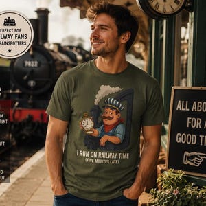 Puede incluir: Camiseta verde oliva con un dibujo de un maquinista de dibujos animados sosteniendo un reloj de bolsillo, con el texto "I RUN ON RAILWAY TIME (FIVE MINUTES LATE)". Un letrero dice "ALL ABOARD FOR A GOOD TIME".