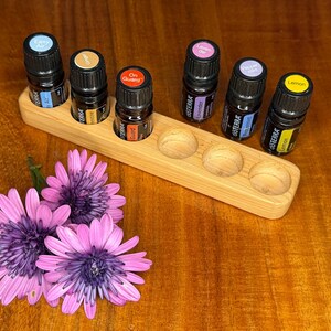 May include: A wooden essential oil holder with six small bottles of essential oils. The bottles have black caps with coloured labels. The holder is rectangular with rounded depressions for the bottles. Pink and purple flowers are in the foreground.