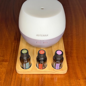 May include: A white and lilac essential oil diffuser with three essential oil bottles on a wooden tray. The bottles are labelled "Eucalyptus", "On Guard", and "Serenity". The diffuser has the "dōTERRA" logo.