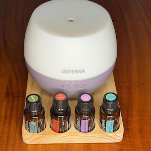 May include: A white and lilac essential oil diffuser with the brand name "dōTERRA" printed on the front. Four essential oil bottles with colourful labels sit in a wooden tray below the diffuser.