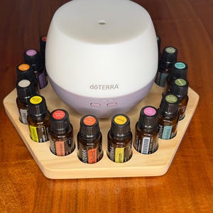 May include: A white and lilac essential oil diffuser, branded "dōTERRA", is encircled by small brown glass bottles of essential oils. The bottles have coloured caps and labels, arranged on a light wood hexagonal tray.