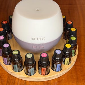 May include: A white and lilac essential oil diffuser, branded with "dōTERRA", surrounded by a wooden tray. The tray holds several small brown glass bottles of essential oils with colourful labels. The diffuser has control buttons.