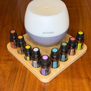 May include: A white and lilac essential oil diffuser, branded "doTERRA", on a wooden tray. It is surrounded by small essential oil bottles with colourful labels. The bottles are filled with essential oils.