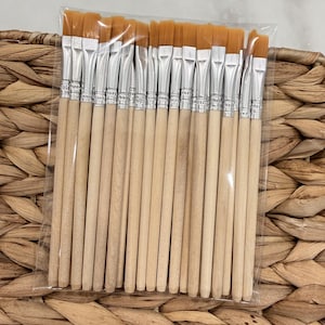May include: A set of paintbrushes with light-colored wooden handles, silver metal ferrules, and flat, golden-brown synthetic bristles. The brushes are arranged side-by-side and enclosed within a clear, rectangular plastic packaging sleeve.