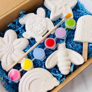 May include: A craft kit with various white ceramic shapes, including a star, turtle, flower, bee, rainbow, and popsicle. The kit includes a paintbrush and small paint pots with various colors, all nestled in a cardboard box with blue paper filler.