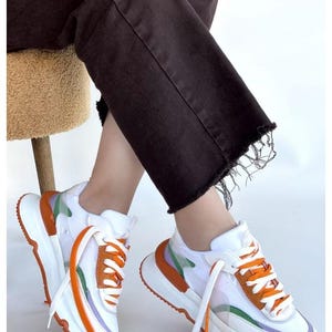May include: White and orange sneakers with a chunky sole. The sneakers feature orange laces, and accents of green and purple. The shoes are worn with dark brown denim jeans.