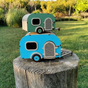 May include: Two stacked wooden camper trailer models. The top trailer is mint green with a cream-colored door and wheel. The bottom trailer is bright blue with a cream-colored door and wheel. Both feature arched doors and rectangular windows.