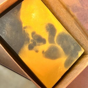 May include: A rectangular bar of handmade soap with a striking color contrast. The soap features a vibrant yellow hue on one side, transitioning to a deep charcoal gray on the other, with dark spots. The soap is presented in a brown box.
