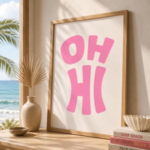 Peut inclure: Une affiche murale encadrée rectangulaire présentant le texte rose OH HI dans une police grasse et arrondie sur un fond crème. Le cadre est en bois clair et l'impression est exposée dans un espace lumineux avec une vue côtière.