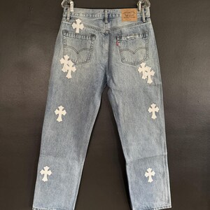 May include: Light blue denim jeans with white cross appliques. The jeans have a classic five-pocket design, a red Levi's tag, and are hanging on a clear plastic hanger. The jeans are a light wash.