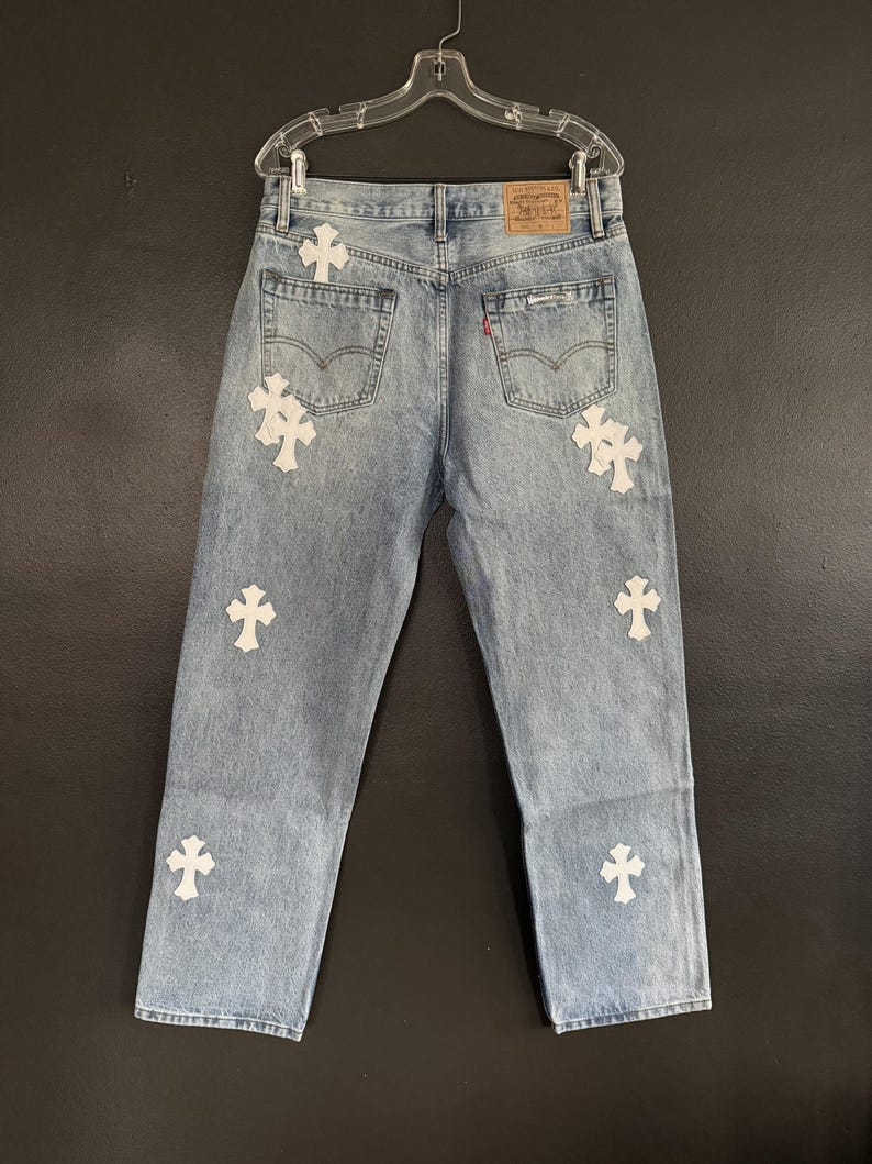 May include: Light blue denim jeans with white cross appliques. The jeans have a classic five-pocket design, a red Levi's tag, and are hanging on a clear plastic hanger. The jeans are a light wash.