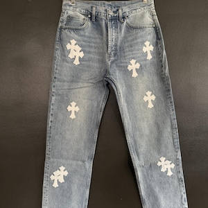 May include: Light wash denim jeans with multiple white cross appliqu&eacute;s. The jeans have a classic five-pocket design and a button and zipper closure. The jeans are hanging on a clear plastic hanger.