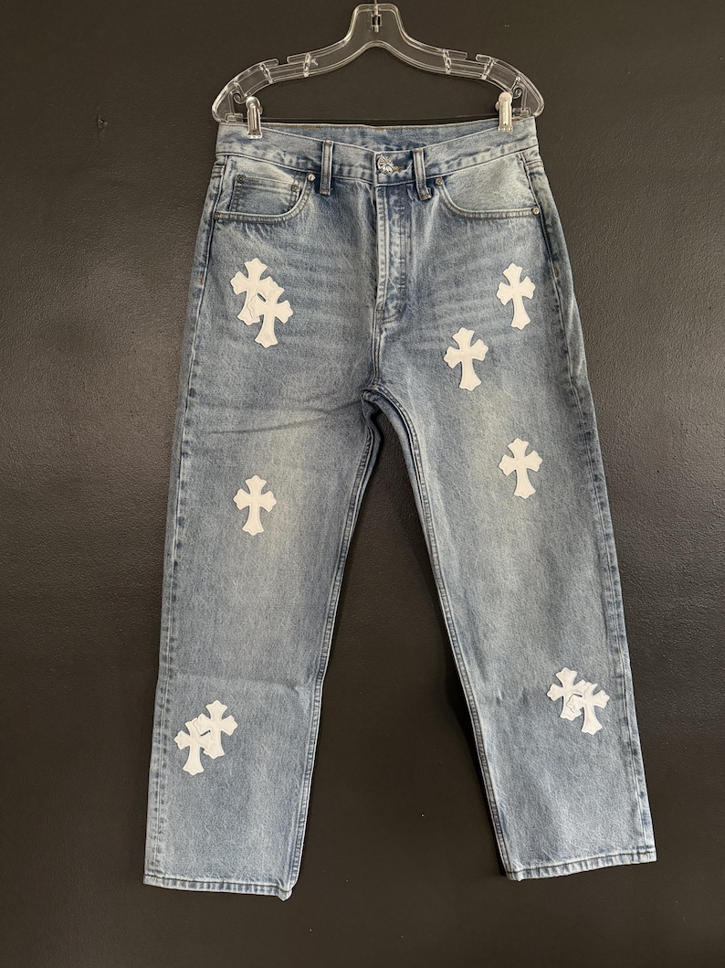 May include: Light wash denim jeans with multiple white cross appliqu&eacute;s. The jeans have a classic five-pocket design and a button and zipper closure. The jeans are hanging on a clear plastic hanger.