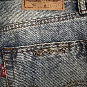 May include: Close-up of blue denim jeans with a brown leather patch and a silver Chrome Hearts emblem on the back pocket. The jeans have a 32-inch waist and a 32-inch inseam. The Levi's red tag is visible.