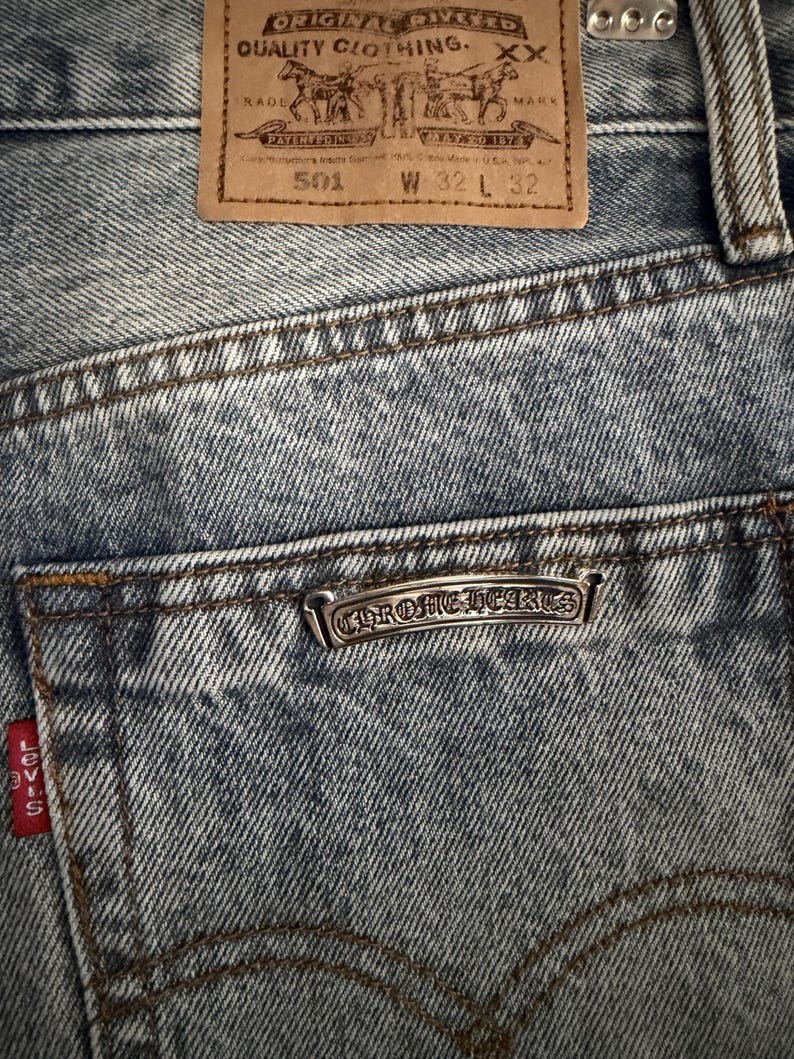 May include: Close-up of blue denim jeans with a brown leather patch and a silver Chrome Hearts emblem on the back pocket. The jeans have a 32-inch waist and a 32-inch inseam. The Levi's red tag is visible.