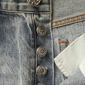 May include: Close-up of light blue denim jeans with silver-colored decorative buttons. The buttons have a floral design. The jeans are slightly faded and show signs of wear.
