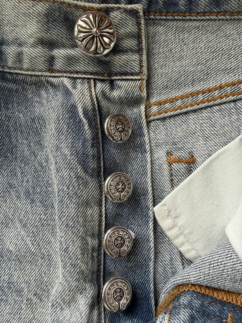 May include: Close-up of light blue denim jeans with silver-colored decorative buttons. The buttons have a floral design. The jeans are slightly faded and show signs of wear.