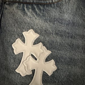 May include: Close-up of blue denim jeans with a pocket. Two white leather cross patches are sewn onto the jeans. The crosses have a decorative edge and are stitched with white thread.