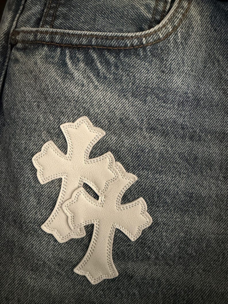 May include: Close-up of blue denim jeans with a pocket. Two white leather cross patches are sewn onto the jeans. The crosses have a decorative edge and are stitched with white thread.