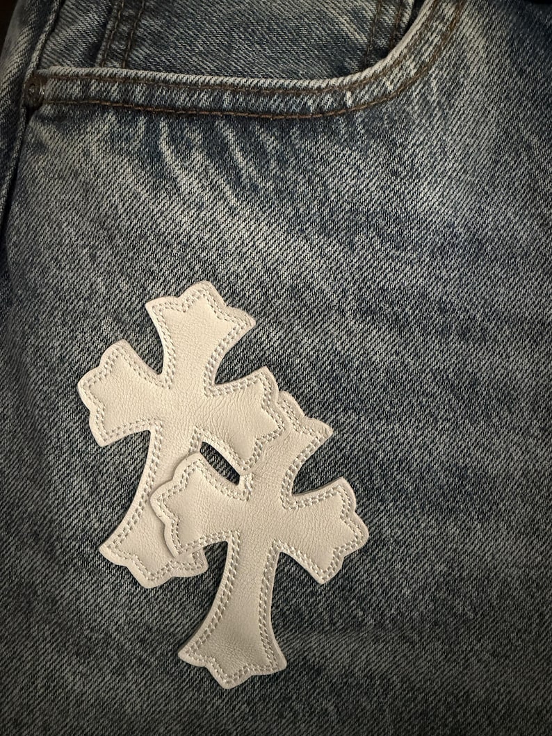 May include: Close-up of blue denim jeans with a pocket. Two white leather cross patches are sewn onto the jeans. The crosses have a decorative edge and are stitched with white thread.
