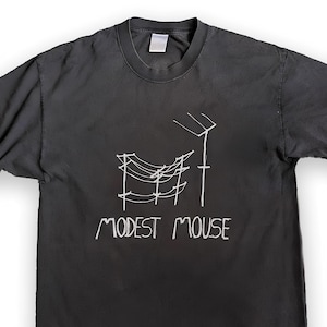 Peut inclure: Un t-shirt noir à manches courtes avec un graphisme en lignes blanches représentant des poteaux électriques avec des câbles et une antenne. Sous le dessin, le texte MODEST MOUSE est imprimé dans une police blanche manuscrite.