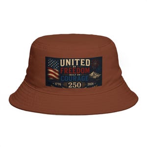 Puede incluir: Un sombrero de pescador marrón con un diseño patriótico. El sombrero presenta un gráfico rectangular con el texto "UNITED BY FREEDOM BUILT ON COURAGE 250" y una bandera estadounidense. Es un accesorio informal.