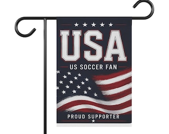 Op de afbeelding: Een rechthoekige tuinvlag hangend aan een zwarte metalen standaard. De vlag heeft een donkerblauwe achtergrond met de witte tekst USA, US SOCCER FAN en PROUD SUPPORTER. Onderaan staat een gestileerde Amerikaanse vlag met rode en witte strepen.