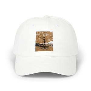 May include: White baseball cap with a curved brim. The cap features a rectangular graphic of a tree with fall foliage and the text "KEEP GOING" and "THE WORK YOU DO TODAY WILL PAY OFF TOMORROW".