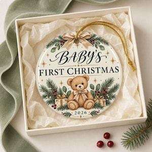 Può includere: Un ornamento rotondo in ceramica bianca con l'illustrazione di un orsacchiotto marrone, rami di pino verdi, bacche rosse e due pacchetti regalo. Il testo riporta "BABY'S FIRST CHRISTMAS" e "2026". In alto è presente un fiocco dorato con cordino.