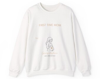 Op de afbeelding: Een wit sweatshirt met lange mouwen en een ronde hals, voorzien van de tekst "FIRST TIME MOM" in beige letters. Onder de tekst staat een lijntekening van een zwanger persoon met een klein beige hartje op de buik, gevolgd door de tekst "Growing a tiny human and a whole new heart."