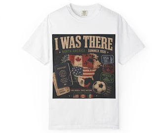 Op de afbeelding: Een wit t-shirt met een grafische print met de tekst I WAS THERE, NORTH AMERICA, SUMMER 2026, ONE MONTH, THREE NATIONS, A LIFETIME MEMORY. Het ontwerp bevat een kaart van Noord-Amerika, een paspoort, een voetbal en diverse reisstempels.
