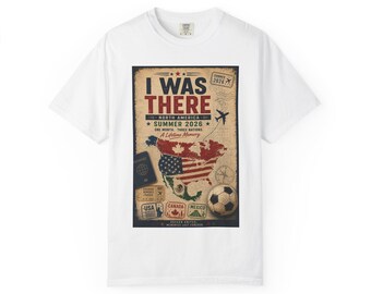 Op de afbeelding: Een wit t-shirt met korte mouwen en een grafische print van een kaart van Noord-Amerika, ingekleurd met de vlaggen van de VS, Canada en Mexico. De tekst luidt I WAS THERE, NORTH AMERICA, SUMMER 2026, A LIFETIME MEMORY en SOCCER UNITED.