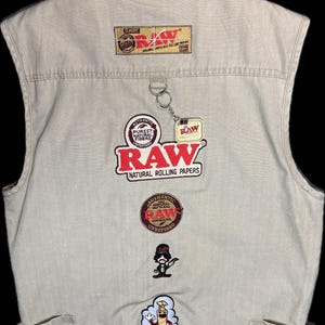 Op de afbeelding: Beige vest met meerdere geborduurde patches en een sleutelhanger. Patches tonen het "RAW" logo, tekst met "PUREST NATURAL FIBERS" en "NATURAL ROLLING PAPERS". Het vest heeft een klassiek ontwerp met zakken en verstelbare zijbanden.
