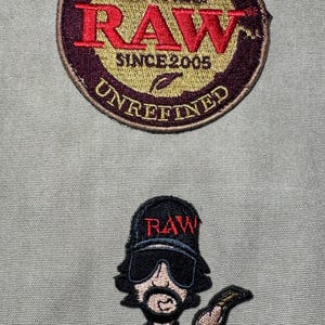 Może przedstawiać: Dwie haftowane naszywki. G&oacute;rna naszywka jest okrągła ze słowami "AUTHENTIC THE NATURAL WAY TO ROLL RAW SINCE 2005 UNREFINED". Dolna naszywka to postać z kresk&oacute;wki w czapce z napisem "RAW".