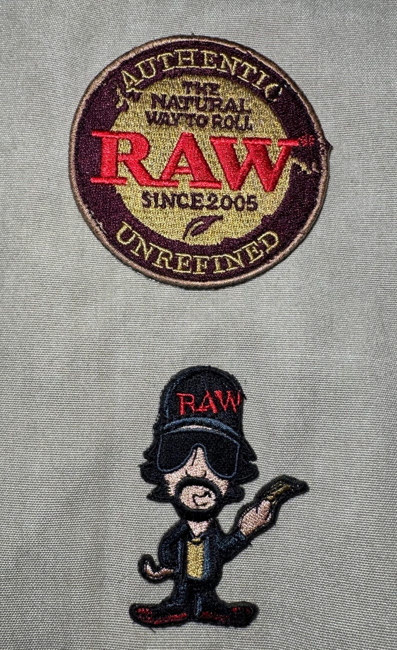 Może przedstawiać: Dwie haftowane naszywki. G&oacute;rna naszywka jest okrągła ze słowami "AUTHENTIC THE NATURAL WAY TO ROLL RAW SINCE 2005 UNREFINED". Dolna naszywka to postać z kresk&oacute;wki w czapce z napisem "RAW".