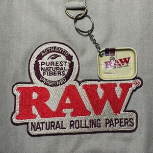 Może przedstawiać: Beżowy materiał z dużym, czerwonym haftowanym logo "RAW", z napisem "NATURAL ROLLING PAPERS" poniżej. Dołączony jest mały metalowy brelok z miniaturową puszką na bibułki "RAW".
