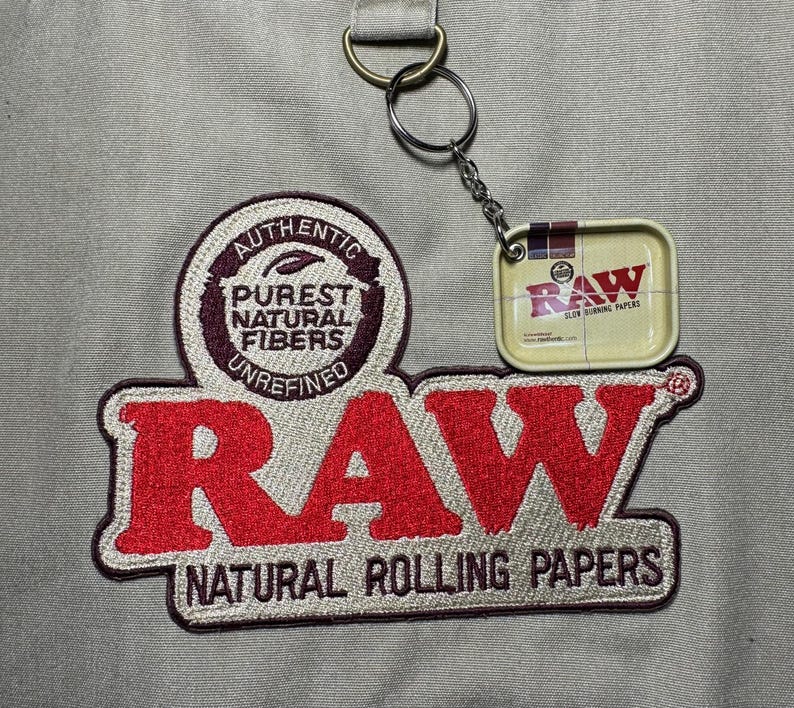 Może przedstawiać: Beżowy materiał z dużym, czerwonym haftowanym logo "RAW", z napisem "NATURAL ROLLING PAPERS" poniżej. Dołączony jest mały metalowy brelok z miniaturową puszką na bibułki "RAW".