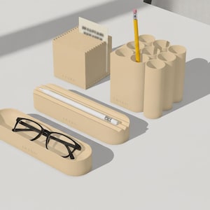 Pode incluir: Um conjunto de acessórios de secretária bege, incluindo um suporte de notas quadrado, um organizador de canetas com vários tubos e um lápis amarelo, um tabuleiro retangular com uma caneta stylus branca e um tabuleiro oval com óculos pretos.