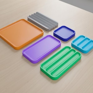 May include: A collection of six small trays in various colours and shapes. The set includes a large orange square tray, a grey ribbed tray, a small blue square tray, a purple rectangular tray, a light blue three-section tray, and a green three-section tray.
