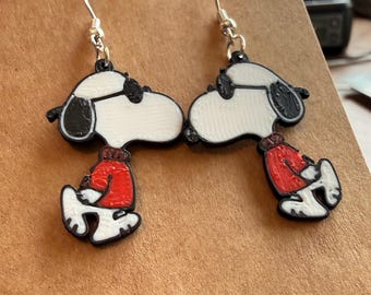 May include: A pair of Snoopy earrings. Each earring features a white Snoopy figure with black accents, wearing a red sweater. The earrings have silver-colored hooks.