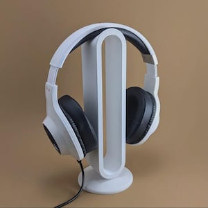 May include: White headphones with black earcups rest on a white headphone stand. The stand has a curved design with a circular base. A black cord extends from the headphones. The background is a solid tan color.