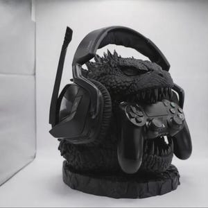 May include: Black gaming headset and controller holder shaped like a Godzilla head. The headset rests on the head, and the controller is held in the mouth. The holder is black and has a textured finish.