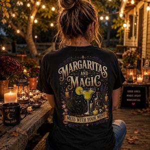 May include: A black t-shirt featuring a graphic print on the back with the text "MARGARITAS AND MAGIC" and "MESS WITH YOUR MIND". The design includes a margarita glass, a black cat, a potion bottle, candles, and floral elements in gold and purple.