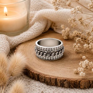 May include: A silver-toned spinner ring featuring a central band of interlocking chain links flanked by two rows of small, clear, round-cut stones. The ring has a polished metallic finish and a wide, flat band design with a smooth interior surface.