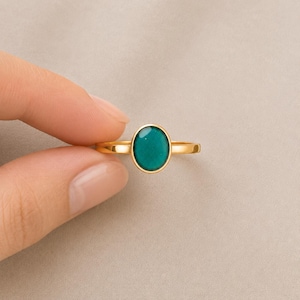 May include: A gold ring featuring a smooth, oval-shaped teal stone set in a simple metal band. The ring has a polished finish and a vibrant, solid-colored gemstone that sits horizontally across the finger, showcasing a clean and balanced design.