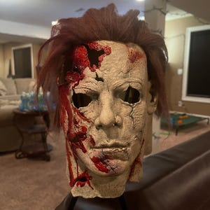 May include: A full-head mask featuring a pale, textured human-like face with dark brown hair. The mask displays significant damage, including deep gashes and holes with bright red blood-like paint splattered across the forehead and cheek.