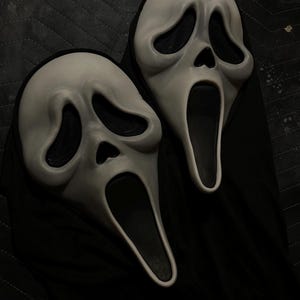 May include: Two white ghost-themed masks with elongated, distorted facial features, including hollow black eye sockets and an open, elongated black mouth. Each mask is attached to a black fabric hood that drapes downward from the base of the face.