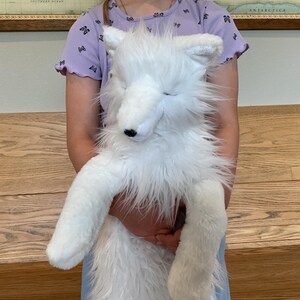 May include: A large, fluffy white plush fox toy with a black nose and closed eyes. The toy has long, soft fur and is being held by a person. The fox has long legs and a tail. The background includes a wooden bench and a map.